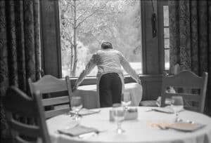 A server lays down a new white tablecloth in a hotel dining room with her back to the camera. She is in front of a window framed by curtains on either side. In the foreground is a table set for a meal.