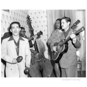 Four men stand in the corner of a house playing instruments. One holds maracas while three play guitars. A bass rests against the wall in the background.