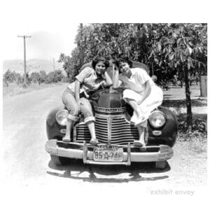 Two young Filipinas sit on the hood of an old car. They are in a rural area with a dirt road and trees.