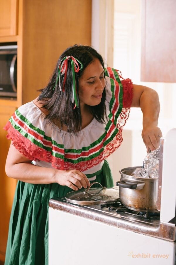 A woman wearing traditional Mexican dress leans over a stove. She is checking on food in a pot on the stovetop.