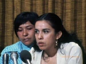 Two women are seated behind microphones. The woman in front is wearing a white jacket.