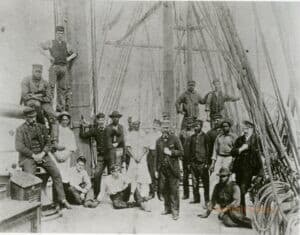 In this old, black-and-white photograph, a number of men stand onboard a ship. The men are are of different ages and races, but primarily white people and Black people.