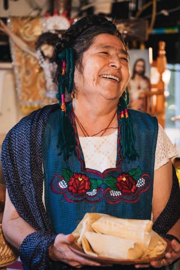 An older woman wearing a blue apron embroidered with flowers holds a plate of tamales. She is smiling and looking to the side of the camera.