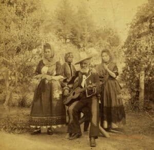 Four people pose in a sepia-tone photograph taken outdoors in 1887. The three people in the foreground - one woman, one older man, and one young woman - are well-dressed. The women stand while the man sits on a stool between them, holding a guitar. The fourth man in the background is looking away from the camera.