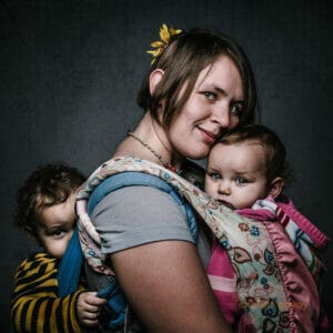 A woman stands with her two children strapped to her front and back. She faces the camera and smiles.