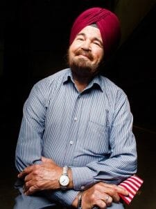 A man in a turban and a blue shirt leans back and smiles at the camera.