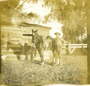 A farm hand stands to the right of a horse and in front of a wood building in a sepia-toned photograph.