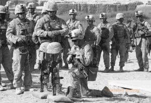 A soldier kneels in front of two rifles standing upright. Helmets are perched on the butt of each rifle. Other soldiers stand nearby.