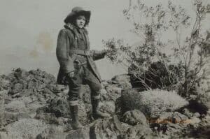 A woman stands on rocks outside. She has a stern expression on her face, wearing men’s clothing, and has a visible sidearm.