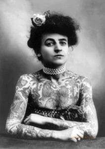 A woman stares defiantly at the camera. She is heavily tattooed and her arms are crossed.