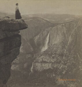 A woman stands at the edge of a cliff in Yosemite National Park in a sepia-toned photograph.
