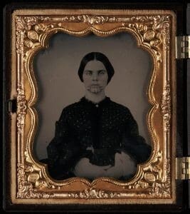 A woman stares straight-faced at the camera. She has lines tattooed on her chin. The photograph is in a gold frame, also seen in the image.