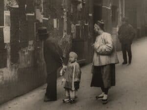 In a sepia-toned photograph, two adults and one child stand on a street. The adults are looking at a bulletin posted on the wall of a building. The child stares straight ahead and holds hands with one adult.
