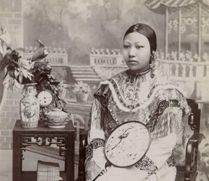 A Chinese woman poses for a portrait in a photography studio in this sepia-toned photograph. The woman is wearing fine clothing and surrounded by Asian-inspired objects, including an intricate table and footstool.