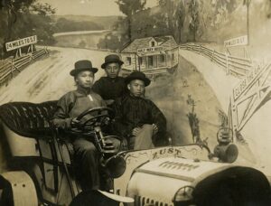 In this sepia-toned photograph, three young Chinese men sit behind the wheel of a car in a photography studio. The backdrop for the photo shows a house and gate. Handwritten on the photograph are the words 