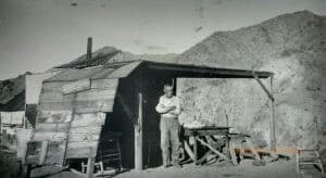 A man stands with his arms crossed outside of a shack with a mountain in the background.