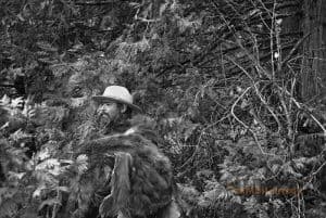 A park ranger stands amongst trees. A bear skin is wrapped around his upper body.