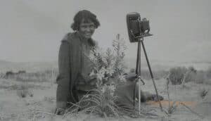A woman sits near a desert lily and old camera in the desert.