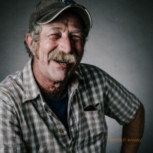 A man with a mustache wears a plaid shirt and baseball cap and faces the camera, smiling.