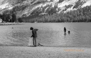 A person holds a garbage bag and picks up trash at the edge of a lake. Mountains are in the background.